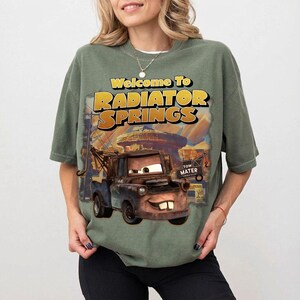 Pu&ograve; includere: T-shirt verde salvia con una grafica di Cricchetto del film Cars, con la scritta "Welcome to Radiator Springs" in giallo e arancione. Il design include un'illustrazione dettagliata del personaggio e della citt&agrave;.
