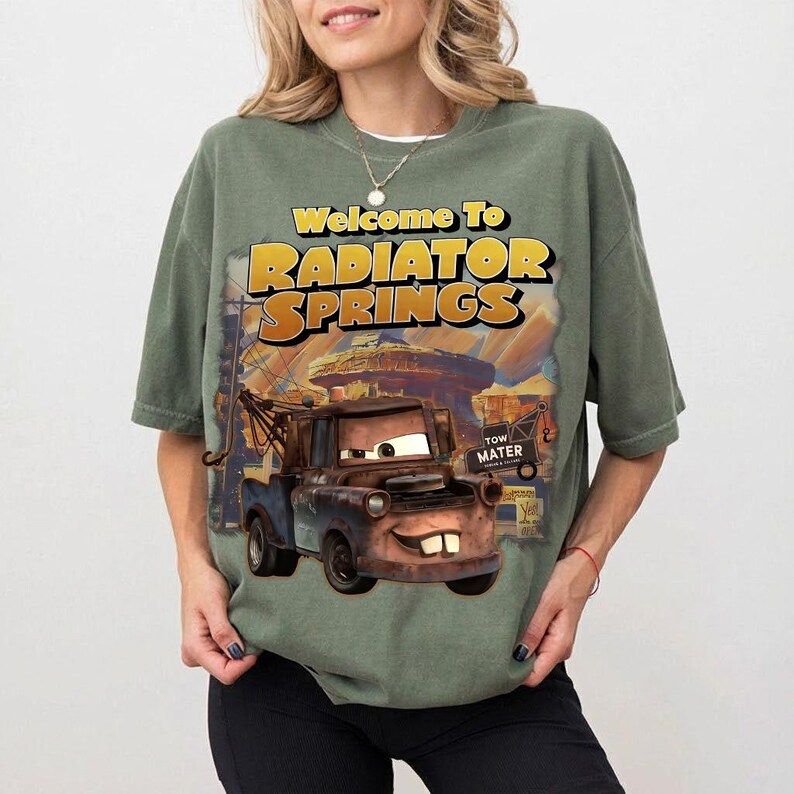 Pu&ograve; includere: T-shirt verde salvia con una grafica di Cricchetto del film Cars, con la scritta "Welcome to Radiator Springs" in giallo e arancione. Il design include un'illustrazione dettagliata del personaggio e della citt&agrave;.
