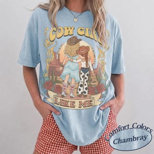 Puede incluir: Una camiseta azul claro con un gr&aacute;fico de dos figuras con sombreros de vaquero en un paisaje des&eacute;rtico con cactus y rosas. El texto dice COW GIRL LIKE ME. La camiseta tiene cuello redondo y mangas cortas, mostrando un ajuste relajado.