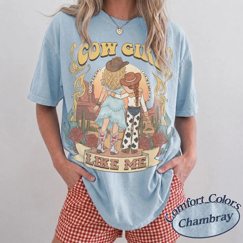 Puede incluir: Una camiseta azul claro con un gr&aacute;fico de dos figuras con sombreros de vaquero en un paisaje des&eacute;rtico con cactus y rosas. El texto dice COW GIRL LIKE ME. La camiseta tiene cuello redondo y mangas cortas, mostrando un ajuste relajado.