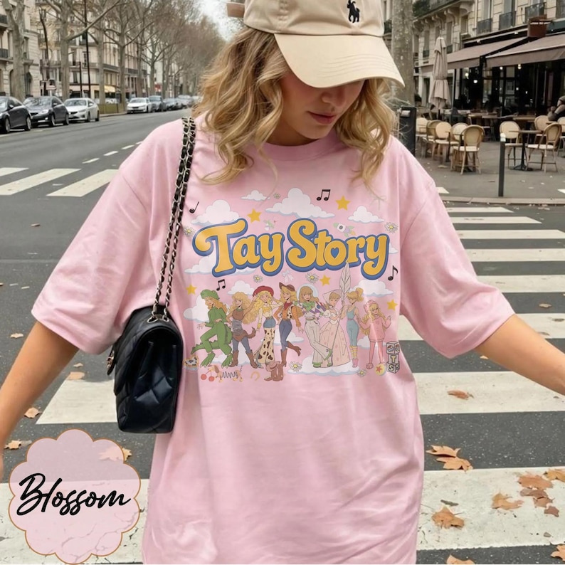 May include: A light pink t-shirt featuring the text Tay Story in yellow and blue letters. The graphic includes a row of illustrated characters in various outfits, surrounded by clouds, stars, and musical notes. A black quilted shoulder bag is worn.