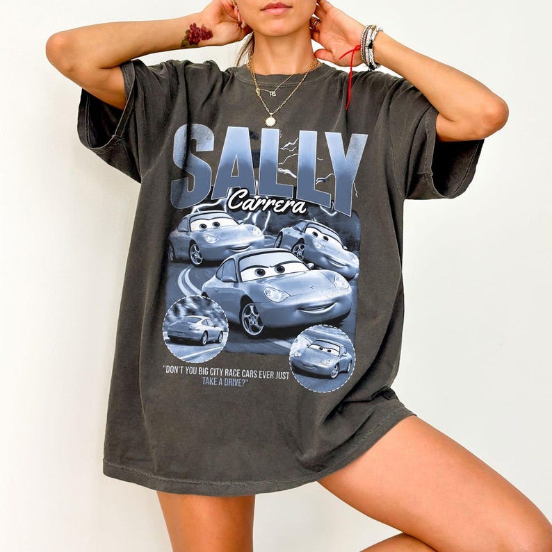 Puede incluir: Camiseta gris oscuro con el texto "SALLY Carrera" y una imagen del personaje Sally Carrera de la pel&iacute;cula Cars. La camiseta tambi&eacute;n incluye la cita: "DON'T YOU BIG CITY RACE CARS EVER JUST TAKE A DRIVE?"