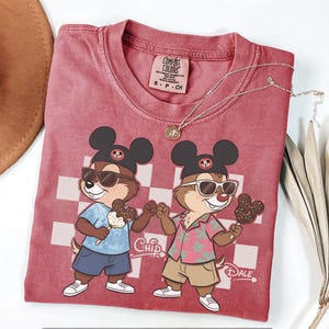 Pu&ograve; includere: Una maglietta Comfort Colors bordeaux con i personaggi di Cip e Ciop che indossano occhiali da sole e tengono in mano gelati a forma di Topolino. La maglietta ha uno sfondo a scacchi e le parole "Chip" e "Dale" sono stampate.