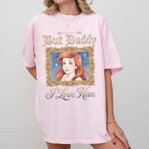 May include: A light pink t-shirt featuring a framed illustration of a red-haired woman with the text "But Daddy I Love Him" and "EST 1909". The shirt has short sleeves and a relaxed fit.