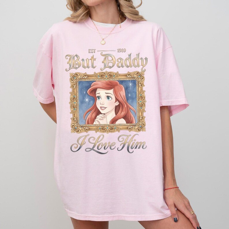 May include: A light pink t-shirt featuring a framed illustration of a red-haired woman with the text "But Daddy I Love Him" and "EST 1909". The shirt has short sleeves and a relaxed fit.