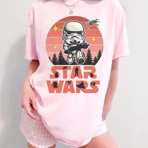 Op de afbeelding: Lichtroze T-shirt met een cartoon Stormtrooper met een blaster, tegen een zonsondergangontwerp met de tekst "STAR WARS". Het shirt bevat silhouetten van Star Wars-personages en -voertuigen.