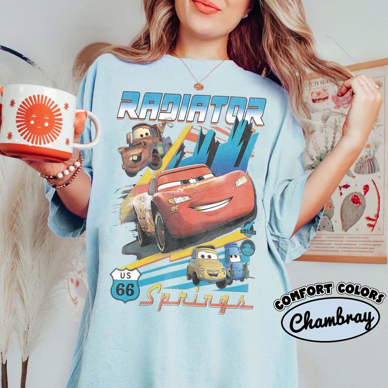 May include: A light blue t-shirt featuring a graphic print of characters from the movie Cars, including Lightning McQueen and Mater, with the text Radiator Springs and US 66. The shirt is held alongside a white mug with a red sun design.