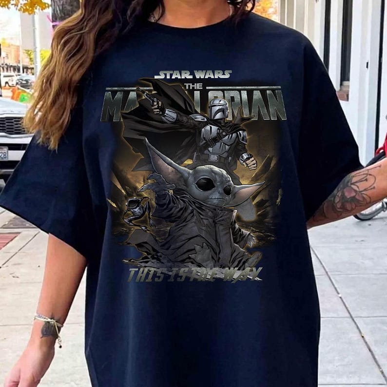 May include: Navy blue t-shirt with a graphic from the Star Wars series, "The Mandalorian." The design includes the text "STAR WARS THE MANDALORIAN" and "THIS IS THE WAY," along with images of the Mandalorian and Grogu.