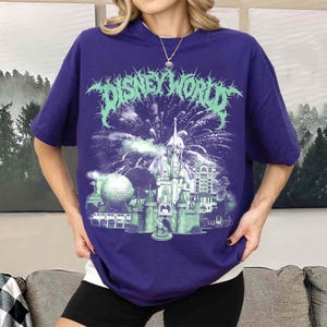 K&ouml;nnte beinhalten: Ein lila T-Shirt mit einer gr&uuml;nen Grafik eines Schlosses, Feuerwerks und dem Text "DISNEY WORLD". Das Design umfasst einen Golfball und andere Geb&auml;ude. Das T-Shirt hat kurze &Auml;rmel.