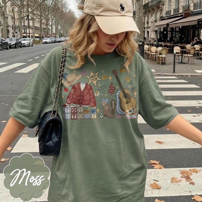 May include: An olive green t-shirt featuring a graphic print of a cowboy hat, a paisley cardigan, a sheriff star labeled Taylor, a microphone, a cowboy boot, a guitar, and blocks spelling Toy Story, paired with a black quilted shoulder bag.