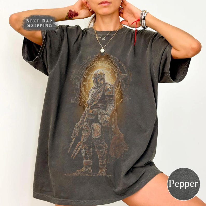 May include: A dark grey t-shirt with a graphic of a Mandalorian warrior. The design features a detailed illustration of the character in armour, set against a circular emblem with text. The shirt has a faded, vintage appearance.