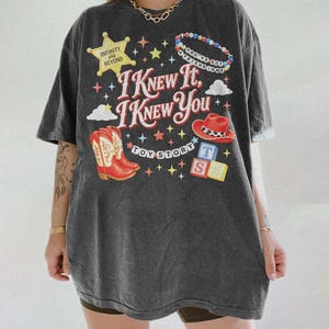May include: A charcoal gray short-sleeve t-shirt featuring a graphic print with the text I KNEW IT, I KNEW YOU, TOY STORY, INFINITY AND BEYOND, and YOU'VE GOT A FRIEND IN ME. The design includes a yellow star, red cowboy boots, a red hat, and blocks.