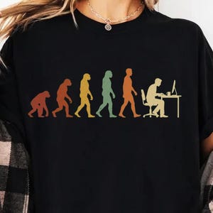 May include: A black t-shirt featuring a multi-colored graphic print that depicts the stages of human evolution, starting from a primate and progressing to a person sitting at a computer desk. The design uses muted tones of orange, yellow, and green.