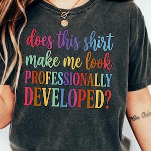 Könnte beinhalten: Ein anthrazitfarbenes Kurzarm-T-Shirt mit dem mehrfarbigen Text "does this shirt make me look PROFESSIONALLY DEVELOPED?" auf der Vorderseite. Das T-Shirt hat einen Rundhalsausschnitt und eine leicht verwaschene, vintage-artige Stoffstruktur.