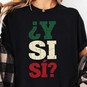 Può includere: Una t-shirt nera a maniche corte con il testo ¿Y SI SÍ? stampato in un carattere usurato e screpolato. Le lettere sono disposte su tre righe, con la riga superiore in verde, quella centrale in crema e quella inferiore in rosso.