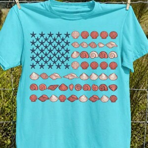 May include: A bright turquoise short-sleeve t-shirt featuring a graphic design arranged like a flag. The left section contains dark blue starfish, while the right section displays rows of various seashells in shades of peach, tan, and coral.