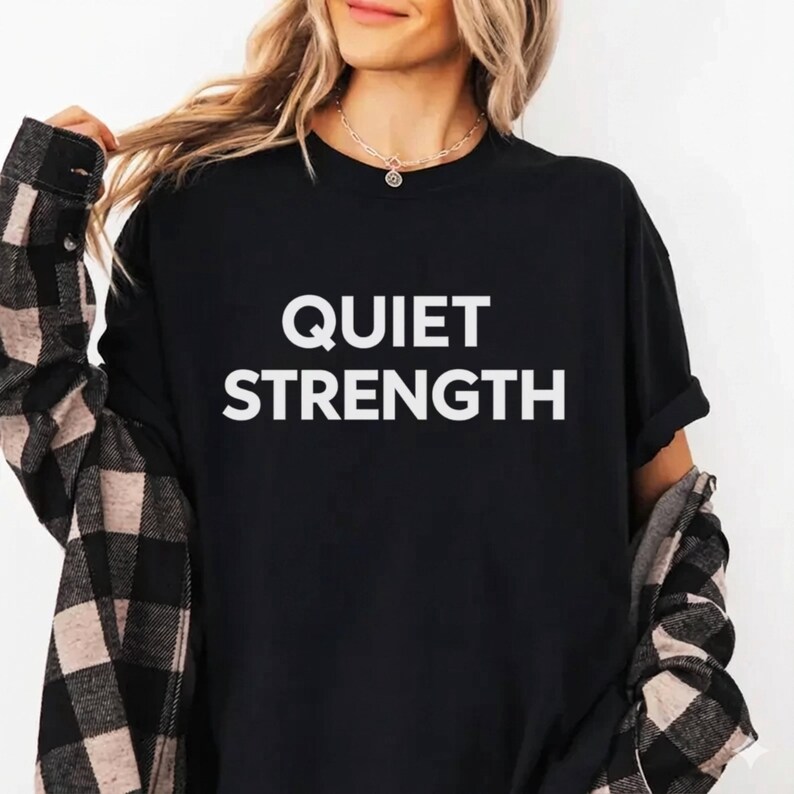K&ouml;nnte beinhalten: Schwarzes T-Shirt mit den Worten "QUIET STRENGTH" in gro&szlig;en, wei&szlig;en Blockbuchstaben. Die Person tr&auml;gt ein schwarz-wei&szlig; kariertes Hemd &uuml;ber dem T-Shirt. Die Person tr&auml;gt auch eine goldene Halskette.
