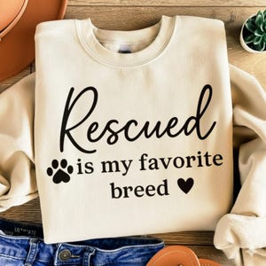 Op de afbeelding: Crèmekleurige sweatshirt met de tekst "Rescued is my favorite breed" in zwart schrift. Een pootafdruk en een hart zijn ook in het ontwerp opgenomen. De sweatshirt ligt op een houten oppervlak.
