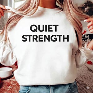 K&ouml;nnte beinhalten: Wei&szlig;es Sweatshirt mit dem Aufdruck "QUIET STRENGTH" in gro&szlig;en, fetten, schwarzen Buchstaben. Das Sweatshirt hat lange &Auml;rmel und einen Rundhalsausschnitt. Das Model tr&auml;gt eine blaue Jeans.