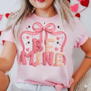 Könnte beinhalten: Hellrosa T-Shirt mit dem gestrickten Schriftzug "BE MINE", verziert mit einer rosa Schleife, Herzen und Sternen. Das Design hat ein Valentinstag-Thema.