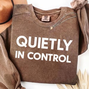 Może przedstawiać: Brązowa bluza Comfort Colors z napisem "QUIETLY IN CONTROL" dużymi białymi literami. Srebrny naszyjnik z małą zawieszką jest zarzucony na koszulę. Koszula jest złożona i umieszczona na białej powierzchni.