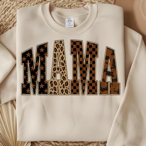 Op de afbeelding: Crèmekleurige sweatshirt met het woord "MAMA" in grote, vette letters. De letters hebben verschillende patronen: sterren, luipaardprint, geblokt en camouflage, allemaal in bruin- en zwarttinten.