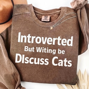 Könnte beinhalten: Ein braunes Comfort Colors Sweatshirt mit dem weißen Text "Introverted But Witing be Discuss Cats". Eine silberne Halskette mit einem kleinen Anhänger liegt auf dem Shirt.