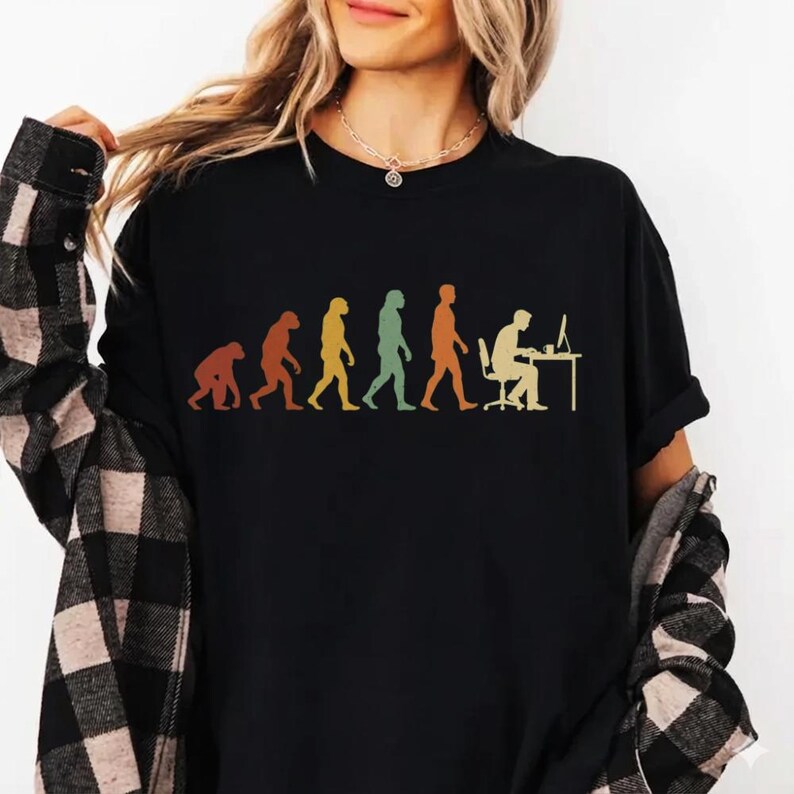 May include: A black t-shirt featuring a multi-colored graphic print that depicts the stages of human evolution, starting from a primate and progressing to a person sitting at a computer desk. The design uses muted tones of orange, yellow, and green.