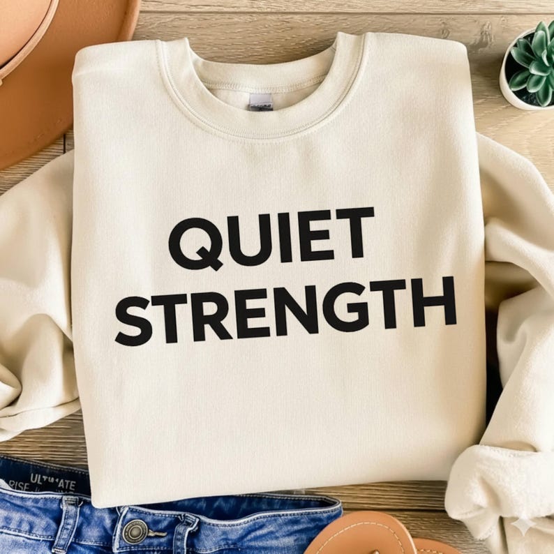 K&ouml;nnte beinhalten: Cremefarbenes Sweatshirt mit dem Aufdruck "QUIET STRENGTH" in fetten schwarzen Buchstaben. Das Sweatshirt liegt auf einer Holzoberfl&auml;che, im Hintergrund sind Jeans und ein Hut zu sehen. Es hat einen Rundhalsausschnitt.
