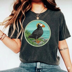 May include: A charcoal gray short-sleeve t-shirt featuring a circular graphic print on the chest. The graphic depicts a black and white puffin with an orange beak standing on a rocky shore with green grass and a blue ocean background.