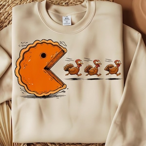 May include: Beige sweatshirt featuring a cartoon pie slice chasing three cartoon turkeys. The pie slice is orange with a bite taken out of it, resembling a Pac-Man character. The turkeys are brown with red waddles and hats, running away from the pie.