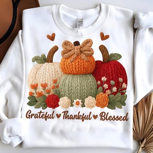 Op de afbeelding: Witte sweatshirt met een ontwerp van drie gestapelde, gebreide pompoenen in oranje, groen en crème, met bloemaccenten en de tekst "Grateful Thankful Blessed" in bruin.