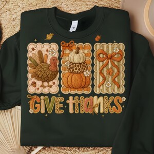 Op de afbeelding: Donkergroene sweatshirt met een Thanksgiving-thema ontwerp. Het ontwerp bevat een kalkoen, gestapelde pompoenen en een grote strik, allemaal binnen decoratieve frames. De zin "GIVE THANKS" wordt weergegeven in een kleurrijk, patroonachtig lettertype.