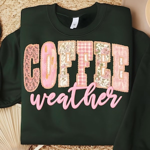 May include: Dark green sweatshirt with the words "COFFEE weather" in a pink and floral design. The word "COFFEE" is in a patchwork style with pink and floral patterns. The word "weather" is in a cursive font.