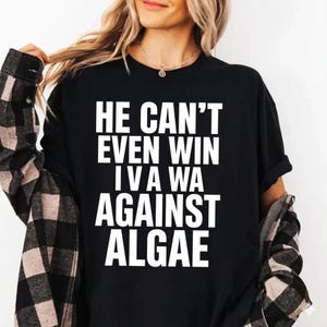 Könnte beinhalten: Ein schwarzes Kurzarm-T-Shirt mit weißem Text, der HE CAN'T EVEN WIN I V A WA AGAINST ALGAE lautet. Das T-Shirt wird unter einem schwarz-weiß karierten Langarmhemd mit Kragen und dunklen Knöpfen an den Manschetten getragen.