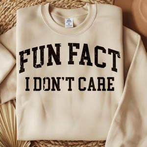 May include: A beige sweatshirt with the text "FUN FACT I DON'T CARE" in distressed black lettering. The crew neck sweatshirt is made of a soft material and is suitable for casual wear.