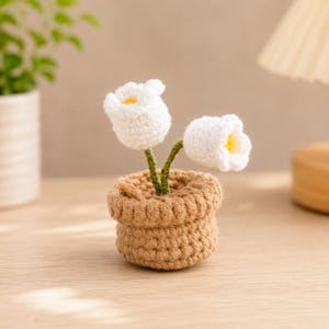 May include: A small crochet potted plant featuring two white flowers with yellow centers and green stems, set in a textured tan yarn pot. The handcrafted item displays intricate stitching throughout the petals and the base, creating a soft, rounded appearance for the decorative piece.