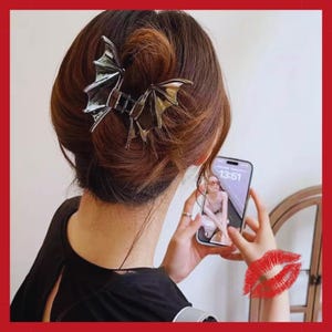 Puede incluir: Una pinza para el pelo plateada con forma de alas de murciélago está sujeta en un moño. La persona sostiene un smartphone que muestra una foto. El fondo es de color claro. Una marca de beso roja está en la esquina inferior derecha.