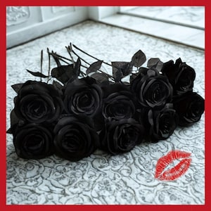 May include: A bouquet of black roses with dark green leaves. The roses are arranged on a white patterned surface. A red lipstick kiss mark is in the lower right corner. The roses are a dark, solid black color.