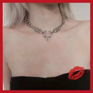 May include: A silver-toned necklace featuring a heart-shaped pendant with flame-like designs. The necklace has a chain-link style and is worn against a black strapless top. A red lip print is visible on the lower right.
