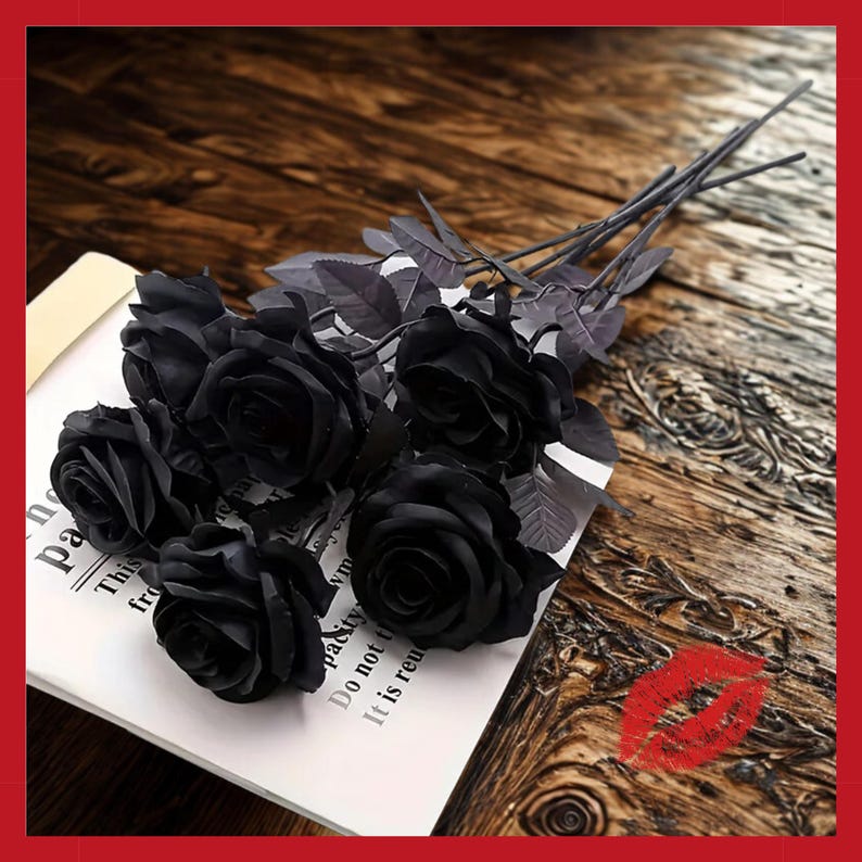 May include: A bouquet of black artificial roses with dark gray stems and leaves. The roses are arranged on a white paper with text, resting on a wooden surface. A red lip print is visible in the corner.