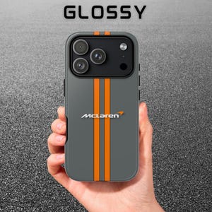Op de afbeelding: Grijze telefoonhoes met oranje racestrepen en het McLaren-logo. Het woord "GLOSSY" staat bovenaan gedrukt. De hoes heeft een glanzende afwerking en is ontworpen voor een smartphone.