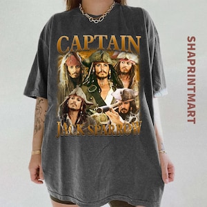 Pode incluir: Camiseta cinza escuro com um gráfico do Capitão Jack Sparrow e outros personagens da série de filmes Piratas do Caribe. O texto "CAPTAIN JACK SPARROW" é impresso em dourado. A camiseta tem mangas curtas.