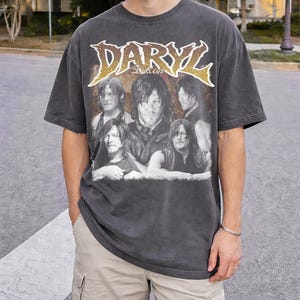 Puede incluir: Camiseta gris oscuro con un gráfico de Daryl Dixon y el texto "DARYL DIXON" en una fuente dorada desgastada. La camiseta tiene un aspecto vintage y cuello redondo.