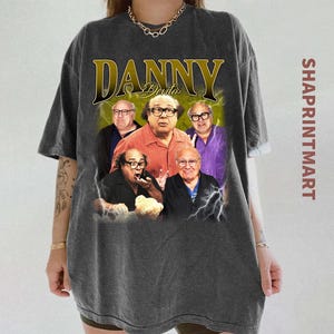 May include: Dark gray t-shirt featuring a graphic of Danny DeVito with the text "DANNY DeVito" in gold. The image includes multiple photos of DeVito with lightning bolt accents. The text "SHAPRINTMART" is printed vertically on the right side.