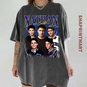 May include: Dark gray t-shirt featuring a graphic of five men with the name "Nathan Scott" in blue script. The design includes a blue background with lightning bolts. The text "SHAPRINTMART" is printed vertically on the right side.