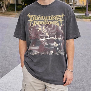 Könnte beinhalten: Dunkelgraues T-Shirt mit einem Fantasy-Motiv mit einem Drachen und einem Krieger sowie dem Text "DUNGEONS & DRAGONS" in Gold. Darunter steht "Never Split the Party". Das Shirt hat kurze Ärmel.