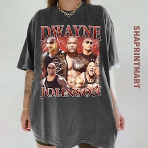 Può includere: T-shirt grigio scuro con una grafica di Dwayne Johnson e la scritta "Dwayne Johnson" in rosso. La maglietta ha un aspetto vintage e la scritta "SHAPRINTMART" è stampata verticalmente sul lato destro.