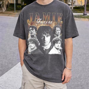 May include: Dark grey t-shirt with the name "Jamie Fraser" in gold lettering and a collage of images. The shirt has a vintage appearance and a crew neck. The images are of a man in different poses.