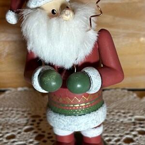 May include: A festive Santa Claus figurine with a red coat, hat, and boots. The figurine has a white beard and holds green mittens. A yellow star on a wire extends from the head. The base is green.